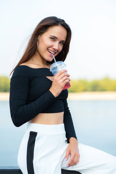Charming brunette girl smiling and drinking cold drink summer outdoor. Young woman's portrait. Female model posing over nature background with river, beach and blue clean sky. - Foto, Imagen
