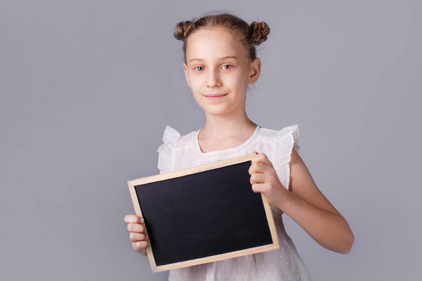 Mała dziewczynka trzyma pustą deskę stojącą na szarym tle. Dziecko pokazuje pustą tablicę. Pojęcie edukacji. - Zdjęcie, obraz
