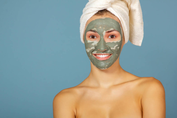 Beautiful cheerful girl applying facial clay mask. Young woman and beauty treatments over blue background. Pampering, youth, anti-aging, body care spa theme. Female face with cosmetics mask - Photo, Image