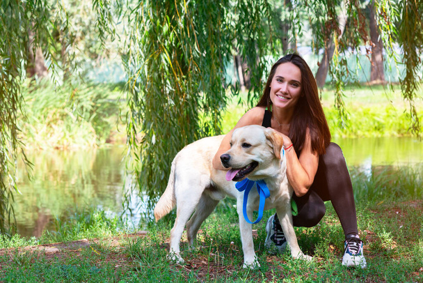 Cheerful pretty young woman sitting and hugging her dog at river bank in summer park - Fotoğraf, Görsel