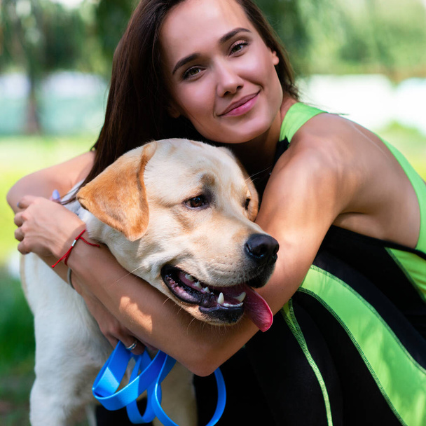 Cheerful pretty young woman sitting and hugging her dog at river bank in summer park - Foto, immagini