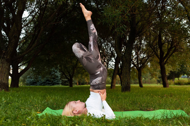 Blonde dans un t-shirt blanc et leggings sportifs faisant du yoga sur un tapis dans le parc
 - Photo, image