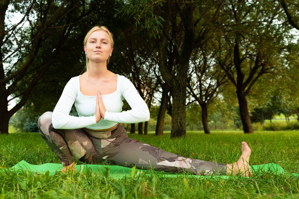 Blondine in weißem T-Shirt und Sportleggings beim Yoga auf einem Teppich im Park - Foto, Bild