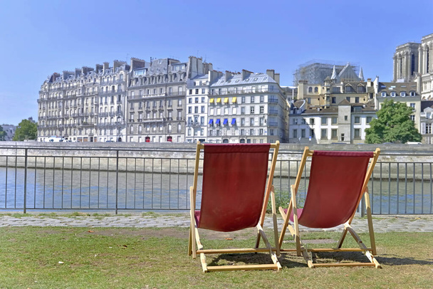 δύο ξαπλώστρες κενές στο γρασίδι στις όχθες του ποταμού Σηκουάνα στο Παρίσι μπροστά από το κτίριο και Notre Dame de Paris  - Φωτογραφία, εικόνα