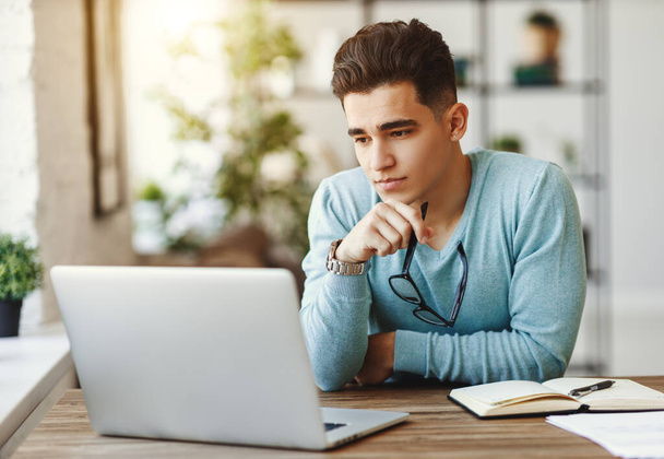 Inteligentny młody mężczyzna w okularach pocierając podbródek i myśląc siedząc przy stole i przeglądając laptop w hom - Zdjęcie, obraz