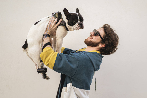 Mladý muž miluje svého psa. Vousáč se slunečními brýlemi drží svého francouzského buldoka čistokrevného psa u bílé zdi venku. Koncept přátelství mužů a psů. - Fotografie, Obrázek