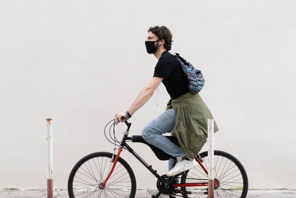 Fiatal felnőtt férfi sétál a városban, biciklizik. Hipster fickó fekete biztonsági maszkot visel egy biciklin egy fehér falnak támasztva. Városi lövés a koronavírus világjárvány idején. - Fotó, kép