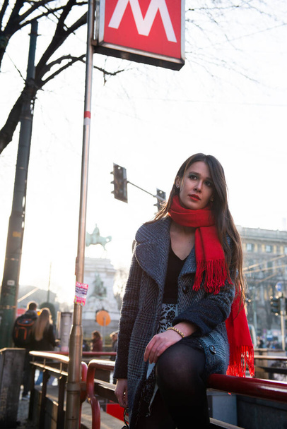 Attractive young woman wearing gray coat with long dark hair red scarf and with sun glasses posing outdoor in Milan streets near metro station, Italy. Beautiful caucasian model portrait, street fashion  - Zdjęcie, obraz