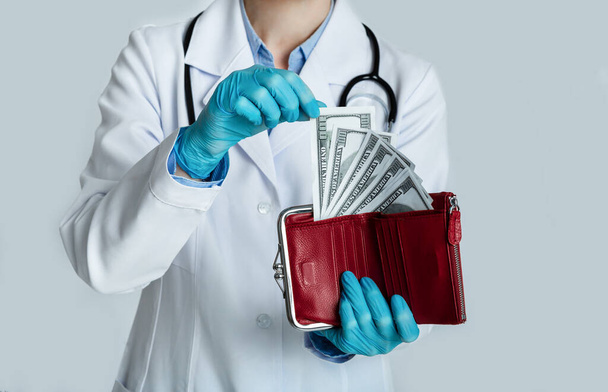 Doctor en bata blanca sostiene monedero rojo con dólares en las manos y toma dinero
 - Foto, imagen