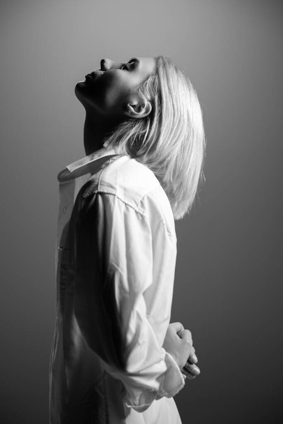 Divatmodell. Egy fiatal szőke nő fehér ingben pózol a stúdióban. Gyönyörű fehér lány szürke háttérrel. Fekete-fehér - Fotó, kép