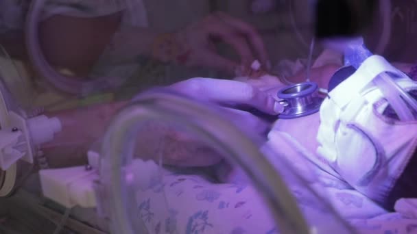 Žena lékař poslouchá srdce dítěte stetoskopem a drží nohu dítěte druhou rukou. Ruce k sobě. Inkubátorové senzory a trubice napojené na kojenecké nohy, ruce a hlavu. - Záběry, video