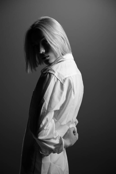 Fashion model. Young blond woman posing in studio wearing white shirt. Beautiful caucasian girl over gray background. Black and white - Photo, Image