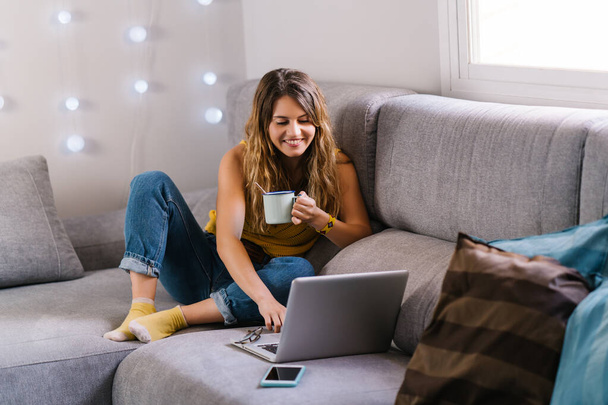 Woman sitting on sofa using laptop and drinking coffee - Foto, Imagen
