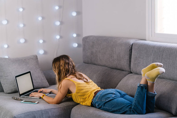 Blonde woman lying on sofa working with laptop at home - Foto, afbeelding