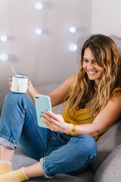Woman sitting on sofa using smart phone and drinking coffee - Fotografie, Obrázek
