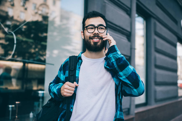 Cheerful bearded asian hipster guy in eyewear for vision correction satisfied with service talking on mobile phone on street, happy millennial male traveler making call in roaming using smartphon - Fotografie, Obrázek