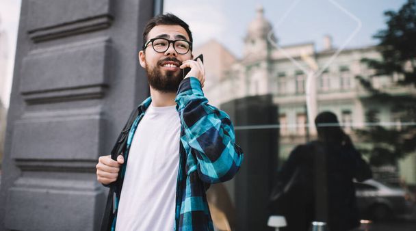 Usmívající se vousatý asijský mladý muž dívá pryč, zatímco se roaming konverzace na mobilní chůze na ulici, pozitivní hipster chlap v ležérní oblečení a brýle mluví na mobilní phon - Fotografie, Obrázek