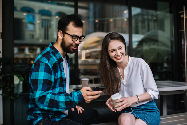 Cheerful multiracial male and female friends having positive conversation discussing ideas during work break in cafe, happy hipster couple drinking coffee to go cup and talking on free time - Foto, immagini