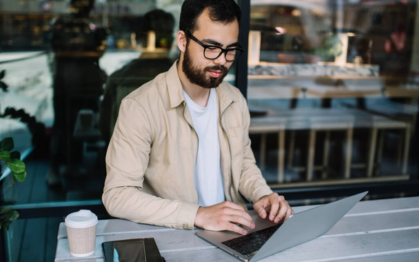 Professional asian bearded it developer working on freelance in eyewear for vision protection typing code on laptop computer in cafe, young hipster guy satisfied with online banking checking app - Фото, изображение
