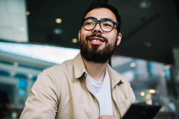 Cheerful bearded asian millennial hipster guy in trendy eyewear and outfit holding mobile phone looking away, happy young handsome man satisfied with 4G connection and smartphone for chattin - Foto, Bild