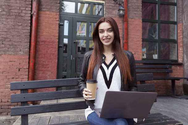 Red-haired girl in a black leather jacket drinks coffee and works at the laptop sitting on a bench in the street - Foto, Imagem