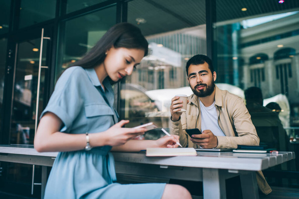 Serious bearded asian hipster guy looking at camera holding cellular while female on frontage checking online information on gadget and making notes, multiracial students using devices in cafe - 写真・画像