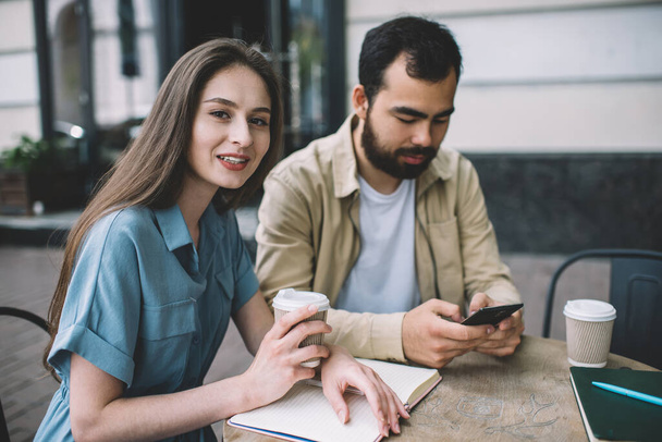 Young attractive caucasian woman looking at camera while her asian boyfriend concentrated on checking smartphone mail spending break in cafe, friends enjoying free time together in coffee shop - Foto, Bild