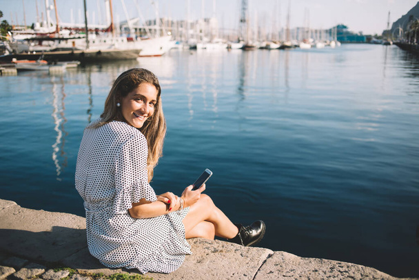 船と港で休んでいる間にカメラで微笑む普段着姿の前向きなヒップスター女の子の肖像,地中海の近くで時間を楽しんで手に携帯電話を持つ幸せな女性観光客 - 写真・画像