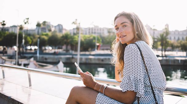 Portrait of attractive hipster girl dressed in stylish apparel looking at camera during leisure and vacations in Spanish city - Barcelona, beautiful female tourist with smartphone device in hand - Photo, Image