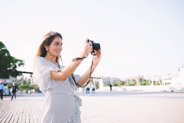 Positive female photographer in stylish dress focusing for clicking good pictures of Barcelona landscape, happy Caucasian woman tourist with modern camera taking pistures during Spanish vacations - Foto, Bild
