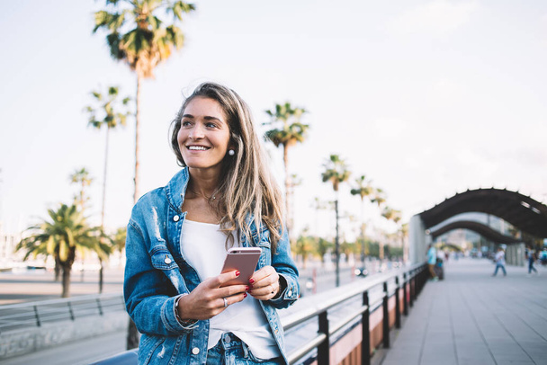 Χαρούμενη γυναίκα blogger ντυμένη με μοντέρνο τζιν μπουφάν χαμογελώντας και κοιτάζοντας μακριά, ενώ περιμένει φιλικό μήνυμα sms στο smartphone gadget, ευτυχισμένη Καυκάσια hipster κορίτσι ξεκουράζεται σε εξωτερικούς χώρους με τηλέφωνο - Φωτογραφία, εικόνα