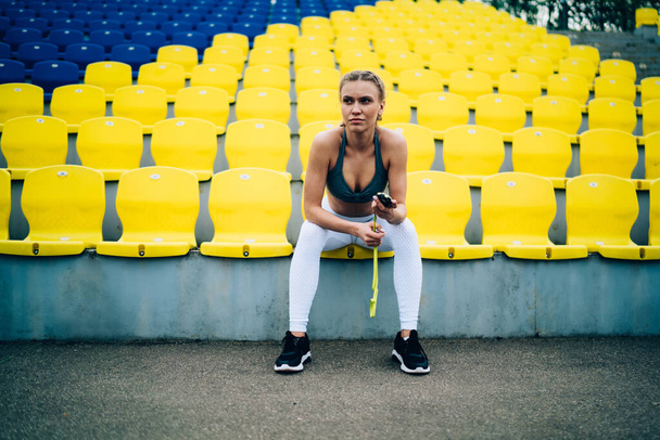 Focused female coach in sportswear with blond pigtails sitting on empty blue and yellow stadium tribunes with stop watch and looking forward - Фото, зображення