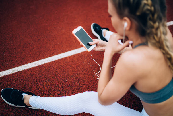 From above adult athletic slim female in modern sportive wear using earphones while using smartphone sitting on ground of track having rest  - Photo, Image