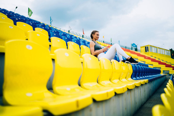 Confident female athlete with blonde hair in white leggings and grey top relaxing with smartphone and looking away on stadium seats - Photo, Image