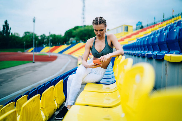 Pensive young slender sportswoman with pigtails in sportswear using smartphone with attention while chilling on tribune on stadium during break - Фото, изображение