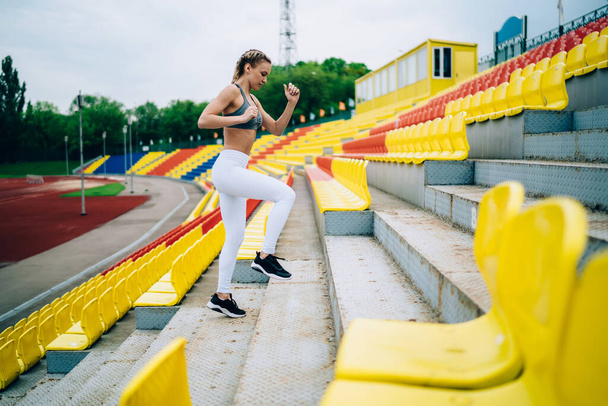 Side view of athletic concentrated sportswoman in sportswear doing stretches for legs jogging on stairs on tribune at stadium looking down - 写真・画像