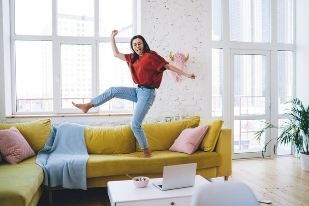 Young smiling woman in casual clothing and glasses dancing on yellow couch in living room near coffee table with laptop and breakfast bowl - Foto, Imagem