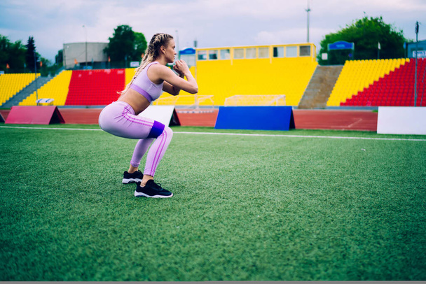 Side view of adult athletic female in modern sportive having training with elastic band squatting on green grass against grandstand of city stadium looking away - Foto, afbeelding