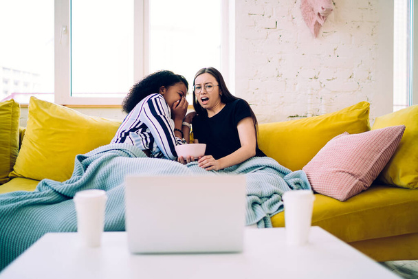 Stunned teen woman with mouth opened and African American girlfriend hiding face while sitting on yellow sofa under blanket and watching horror movie on laptop - Foto, Bild