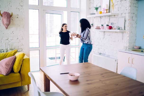 Amused multiracial women in casual modern clothes having conversation and carrying takeaway cups while entering doorway into modern loft apartment - Fotoğraf, Görsel