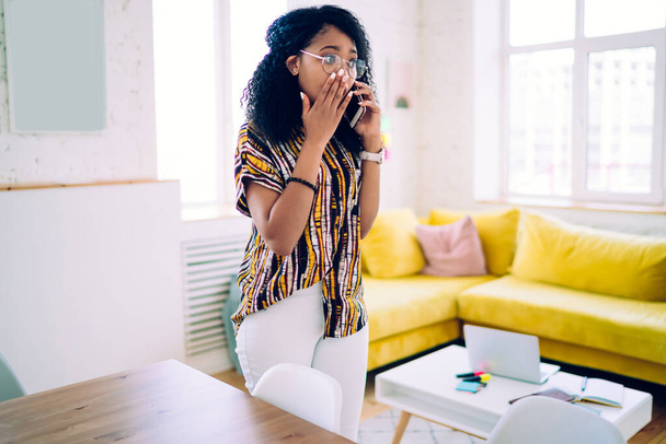 Frustrated African American young woman in domestic clothes and eyeglasses chatting on smartphone and covering mouth while standing at stylish apartment - Fotoğraf, Görsel