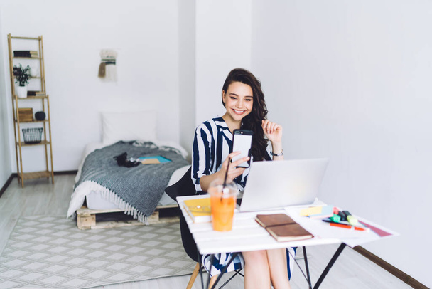 Young happy attractive dark haired woman in casual clothes smiling and taking selfie on smartphone while sitting at desk with laptop in bedroom - Foto, Imagen