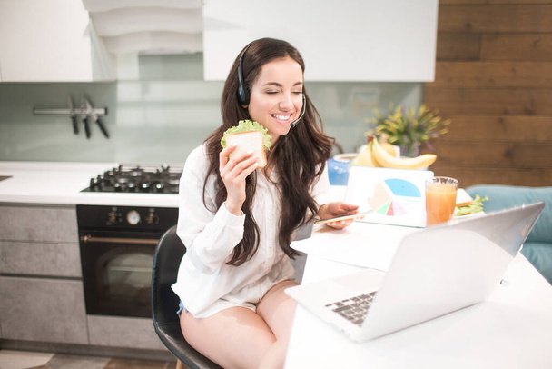 Beautiful black-haired woman works from home and uses headphones with a headset. An employee sits in the kitchen and has a lot of work on a laptop and tablet and has video conferencing and meetings. - Photo, Image