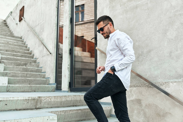 Handsome young bearded man in stylish casual outfit and black sunglasses, goes up the concrete stairs in urban city center outdoors. Hipster male is walking the street for a co-meeting with partners. - Foto, imagen