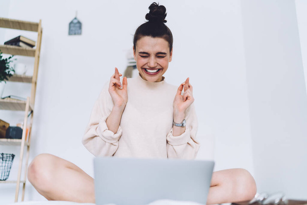 Nenucená mladá žena v pouličním oblečení s širokým úsměvem sedící se zkříženýma nohama a prsty a zavřenýma očima s laptopem na bílém podkrovním pokoji  - Fotografie, Obrázek