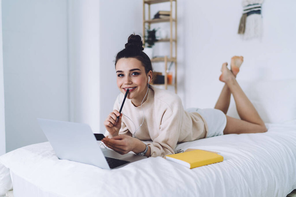 Cheerful pretty woman with hair bun and earphones relaxing on bed holding pen in mouth browsing laptop and looking at camera at home - Zdjęcie, obraz