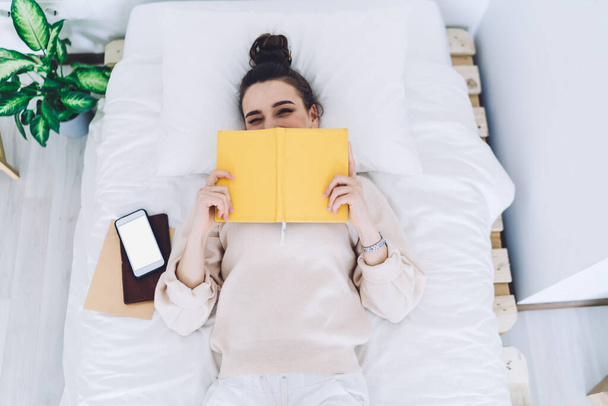 From above laughing charming woman closing half face with open yellow book while lying in bed with white linen and smartphone  - Foto, imagen