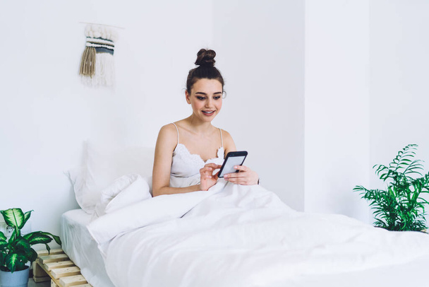 Concentrated smiling female waking up on modern soft bed with white blanket using smartphone in light loft with minimalist interior - Φωτογραφία, εικόνα