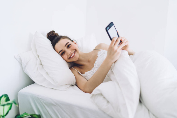 Smiling brunette in nightwear chilling under blanket on comfy bed while taking selfie and looking at camera on white background - 写真・画像
