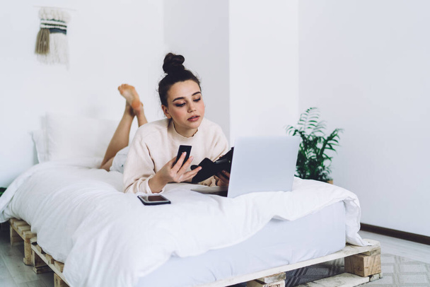 Beautiful brunette in home clothing taking credit card from wallet while lying on handmade bed in room with white interior - Valokuva, kuva